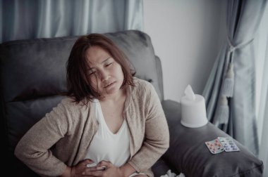 Asyalı hasta kadın kanepede oturur, evde kalır, kadın kendini kötü hisseder, uzanır ve dinlenmek ister, karın ağrısı, regl sancısı...