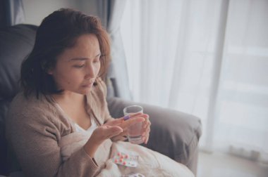 Asyalı hasta kadın kanepede oturur, evde kalır, kadın kendini kötü hisseder, uzanıp dinlenmek ister, ilaç alır.