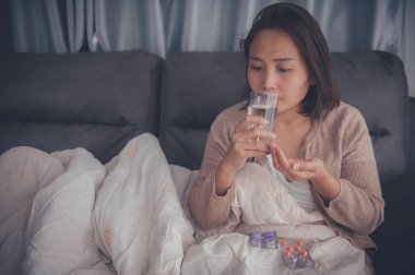 Asyalı hasta kadın kanepede oturur, evde kalır, kadın kendini kötü hisseder, uzanıp dinlenmek ister, ilaç alır.