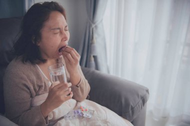 Asyalı hasta kadın kanepede oturur, evde kalır, kadın kendini kötü hisseder, uzanıp dinlenmek ister, ilaç alır.