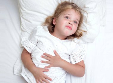 Child girl in bed with hands on her stomach. Caucasian kid with ache.Nausea and vomiting.Sickness.Little girl suffers from desease.
