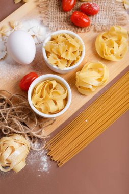 Kahverengi arka planda kuru İtalyan makarnası ve kiraz domatesleri. Makarna geleneksel İtalyan yemeği spagetti, fettucine, farfalle, kiraz domatesleri, yumurta, un renginde ahşap masa üzerindedir. Çiğ İtalyan makarnası, glütensiz.. 