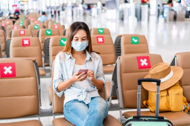 Havaalanında uçuş beklerken ameliyat maskesi takan ve cep telefonu kullanan genç bir kadın. Yeni normal yaşam tarzı kavramı.