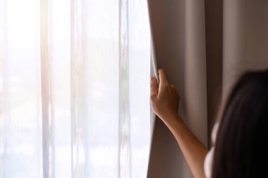 Genç Asyalı kadın sabah perdeleri açıyor ve evdeki pencereden bakıyor.