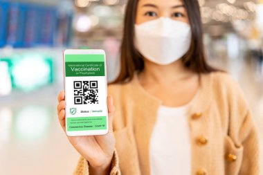 Akıllı telefon tutan ve havaalanında COVID-19 aşı pasaportu gösteren genç bir kadın gezgin.