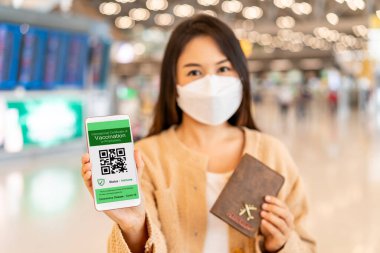 Akıllı telefon tutan ve havaalanında COVID-19 aşı pasaportu gösteren genç bir kadın gezgin.