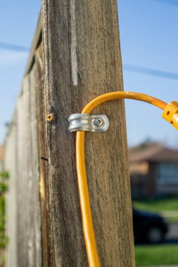 Yellow extension cable through metal bracket screwed to the support pole of a wooden fence. Blurred fence and house in the background