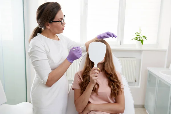 Kozmetológus használ arc marker a női beteg arcát, mielőtt kozmetikai kezelés spa szalonban. Az ügyfél tükörbe néz a szépségklinikán. — Stock Fotó