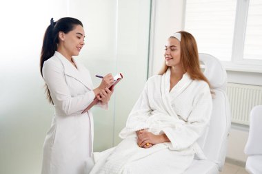 Kozmetik uzmanı kadın hastaya danışmanlık yapıyor ve modern güzellik kliniğinde tıbbi randevu sırasında notlar alıyor.