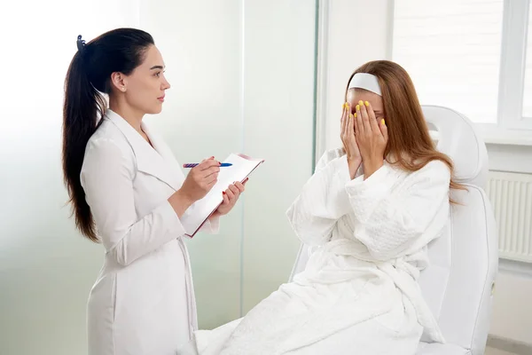 Cosmetólogo consultando paciente mujer asustada y tomando notas durante cita médica en clínica de belleza moderna — Foto de Stock