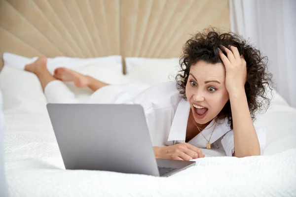 Curly girl using laptop computer sitting on white bed browsing or chatting with friends online Royalty Free Stock Images
