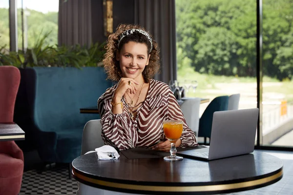 Schöne junge lockige Frau sitzt in einem Café und trinkt frischen Orangensaft und wartet auf ihren Freund — Stockfoto