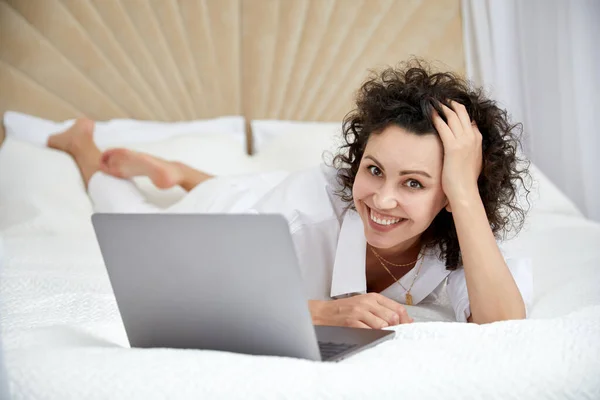 Lockig flicka med hjälp av bärbar dator som sitter på vit säng surfa eller chatta med vänner online — Stockfoto