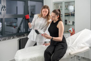 Kozmetolog cilt bakımı için bir paket tutuyor. Ofisinde bir kadın doktor müşteri tedavi seçeneklerini gösteriyor.