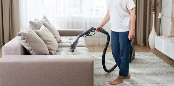 Junge Frau in weißem Hemd und Jeans putzt Teppich unter Sofa mit Staubsauger im Wohnzimmer, Kopierraum. Haushalts-, Reinigungs- und Hauswirtschaftskonzept — Stockfoto