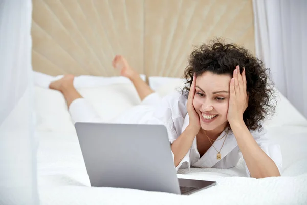 Lockig flicka med hjälp av bärbar dator som sitter på vit säng surfa eller chatta med vänner online — Stockfoto