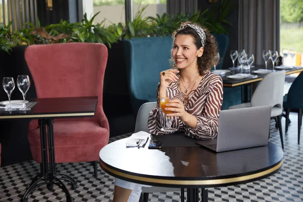 Hermosa mujer rizada joven sentada en un café bebiendo jugo de naranja fresco y esperando a su amigo —  Fotos de Stock