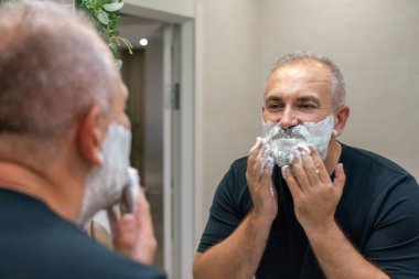 Sakalını ve bıyığını jiletle kesmek için yüzüne tıraş köpüğü süren olgun, yakışıklı bir adam.