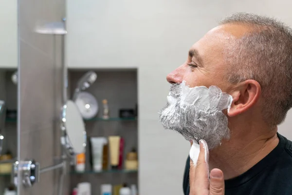Mature handsome man applying shaving foam on his face to cut off his beard and mustache with razor — Stock Photo, Image