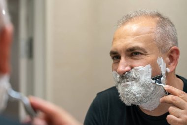 Olgun, kır saçlı bir adam tıraş bıçağı kullanarak sakalını evde dinlendiriyor.