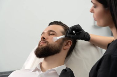 Genç sakallı adam bir güzellik kliniğinde profesyonel kozmetik uzmanı tarafından yüz bakımı yaptırılıyor. Sağlıklı adam yaşam tarzı konsepti