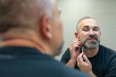 Çekici orta yaşlı bir adam sakalını ve bıyığını aynanın önünde traş ediyor.