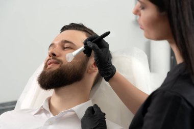 Genç sakallı adam bir güzellik kliniğinde profesyonel kozmetik uzmanı tarafından yüz bakımı yaptırılıyor. Sağlıklı adam yaşam tarzı konsepti