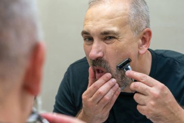 Olgun, kır saçlı bir adam tıraş bıçağı kullanarak sakalını evde dinlendiriyor.