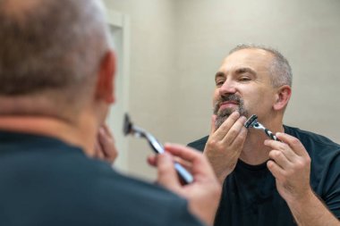 Çekici orta yaşlı bir adam sakalını ve bıyığını aynanın önünde traş ediyor.