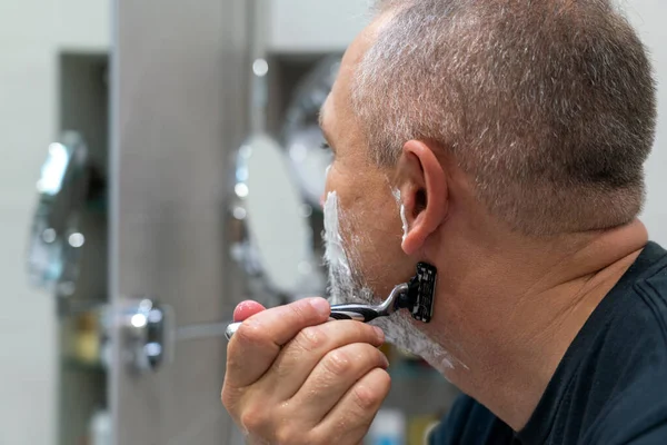 Olgun, kır saçlı bir adam tıraş bıçağı kullanarak sakalını evde dinlendiriyor.