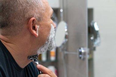 Orta yaşlı yakışıklı adam sakalını ve bıyığını kesiyor banyoda dikilip aynaya bakıyor.