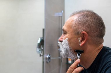 Orta yaşlı yakışıklı adam sakalını ve bıyığını kesiyor banyoda dikilip aynaya bakıyor.