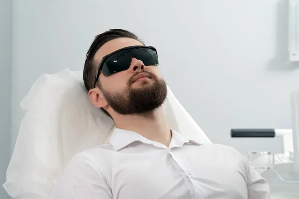 Bearded man sitter innan laseransiktsbehandling av professionell kosmetolog på en skönhetsklinik. Friska man livsstil koncept Royaltyfria Stockbilder