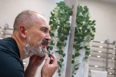 Orta yaşlı yakışıklı adam banyoda sakalını kesiyor.