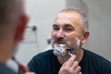 Orta yaşlı yakışıklı adam banyoda sakalını kesiyor.