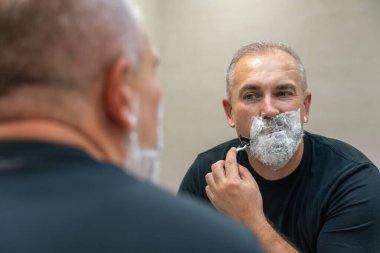 Olgun, kır saçlı bir adam tıraş bıçağı kullanarak sakalını evde dinlendiriyor.