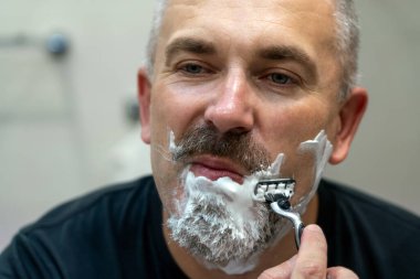 Orta yaşlı yakışıklı adam banyoda sakalını kesiyor.