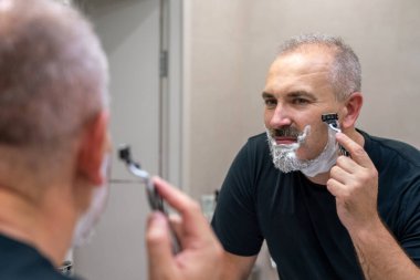 Orta yaşlı yakışıklı adam banyoda sakalını kesiyor.