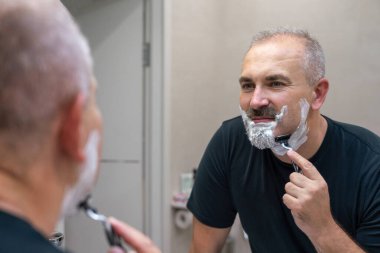 Orta yaşlı yakışıklı adam banyoda sakalını kesiyor.