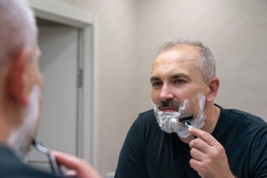 Orta yaşlı yakışıklı adam banyoda sakalını kesiyor.