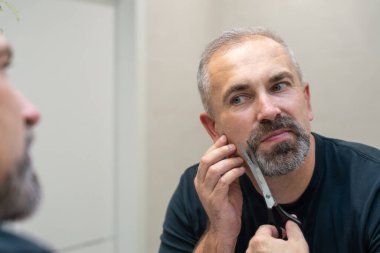 Middle-aged handsome man using scissors to cut his beard a litlie 