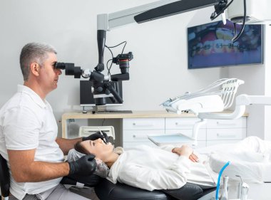 Male dentist using dental microscope treating patient teeth at dental clinic office. Medicine, dentistry and health care concept