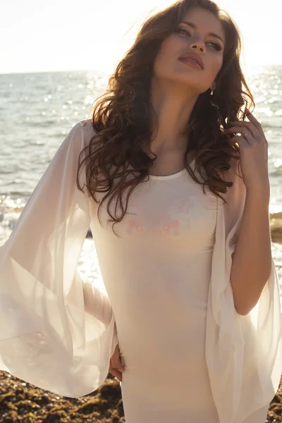 Красивая девушка в белом платье позирует на пляже в солнечных лучах — стоковое фото