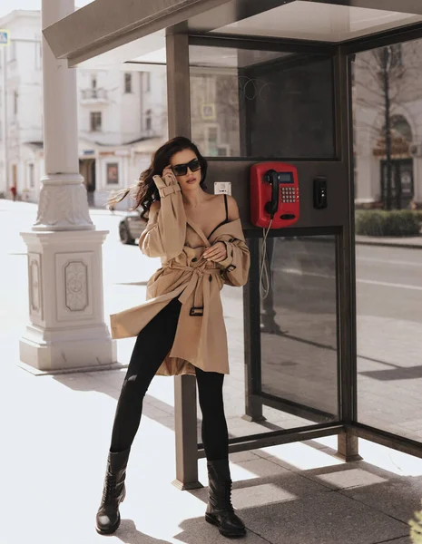 时尚的户外照片 在城市街道上的电话亭里 穿着雅致的米色外套和靴子 一头乌黑的头发 站在美丽的女人面前 — 图库照片