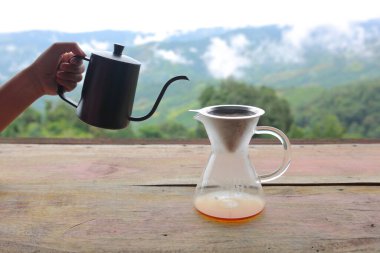 Kettle 'dan damlayan kahve poşeti doğadaki ahşap masa arkasında, sabah kahve saati dağ manzaralı ve doğal yeşil manzaralı soğuk ve rahat..