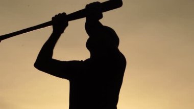 Erkek silueti gün batımına karşı beyzbol sopası tutuyor.
