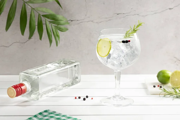 Cold glass of gin tonic and bottle on white wooden base on light background.