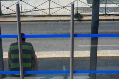 Üzgün adam güneşli bir günde mavi soyulmuş otobüs durağında oturuyor.