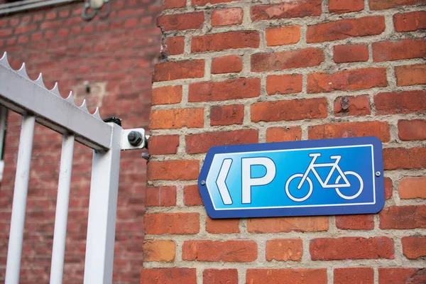 Beyaz çitin yanındaki kırmızı kahverengi tuğla duvarda mavi bisiklet parkı tabelası.