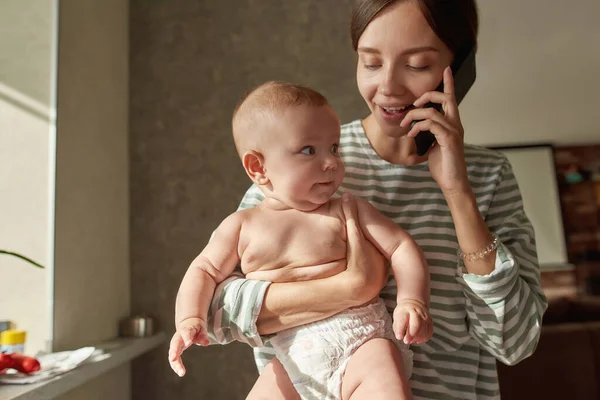 Ibu muda yang ceria berbicara di telepon sambil menggendong bayi — Stok Foto